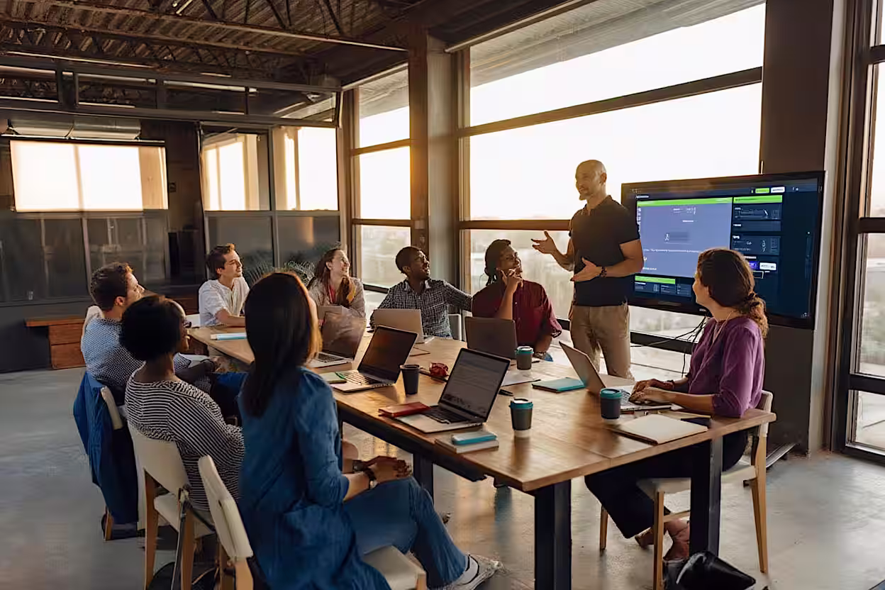 Cyber security meeting with diverse colleagues gathered around a table, with fractional executive standing and presenting using a large screen.