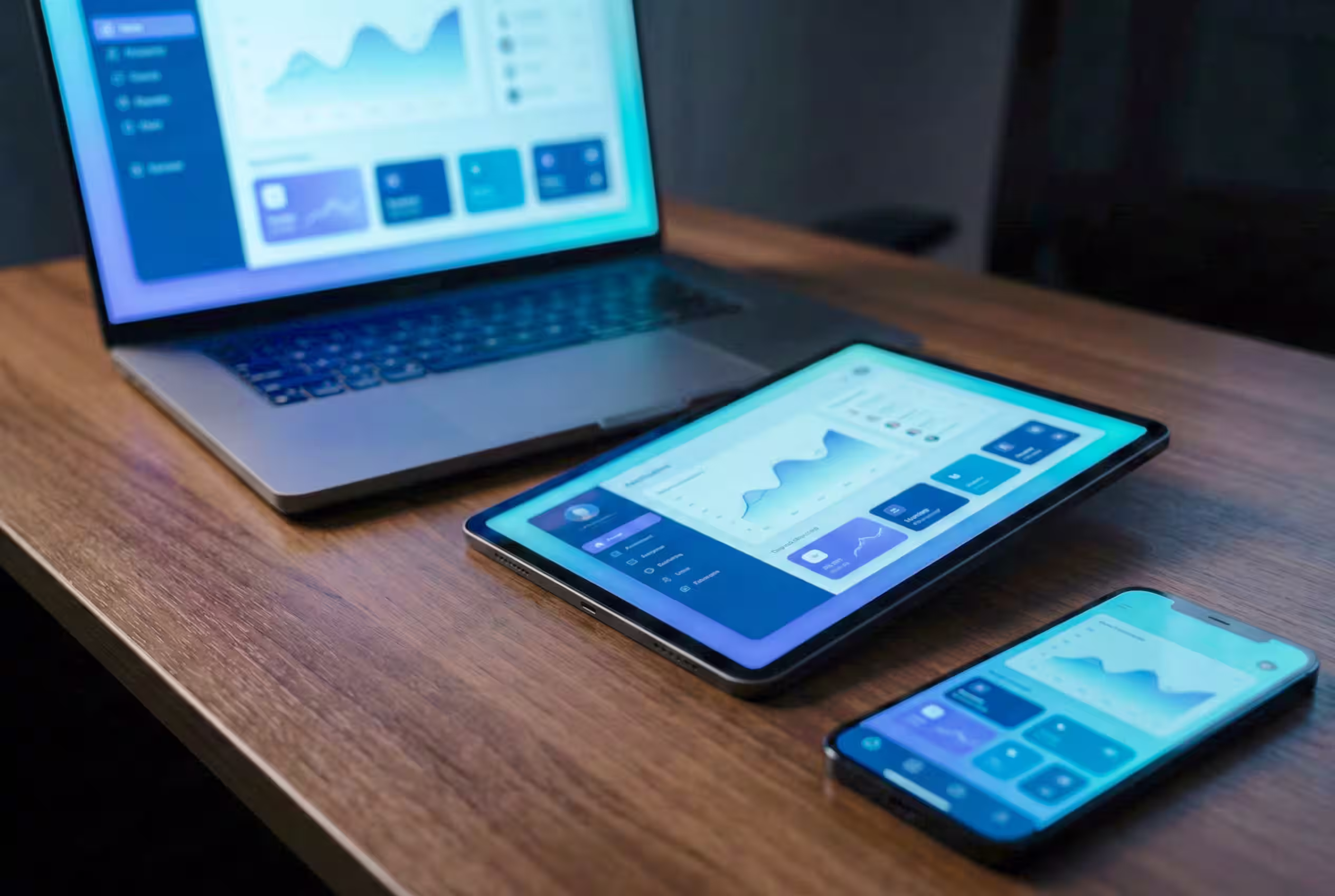 Laptop, tablet, and smartphone on a wooden desk displaying no code application data charts on their screens built with no code solutions like Webflow, Airtable, Outseta and Make.